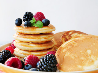 Mini pancakes with raspberries, blueberries and syrup on white plate. Stack of silver dollar pancakes closeup.