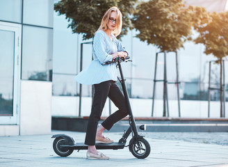 Blond pensive woman in glasses is driving her new electrical scooter on the street at bright sunny day. © Fxquadro