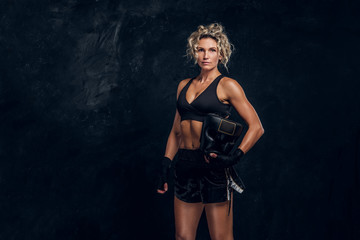 Fototapeta na wymiar Experienced female boxer is posing for photographer at dark photo studio with equipment in her hands.
