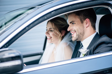 selective focus of attractive bride in bridal veil and bridegroom smiling in car
