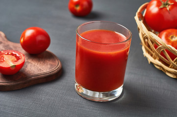 Full glass of fresh juice near wiker wooden basket full of ripe tomatoes and cutting board lies on dark concrete