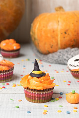 Festive Halloween cupcakes decorated colored decorations witch hat, orange cream, the face of a ghost. Wooden background with pumpkin and copy space, selective focus.