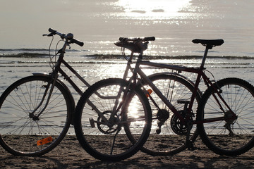 Two bicycles  silhouettes standing on the sand near the water edge on the beach.  Beautiful  shiny and glow water of the sea on the background. Sport and recreation concept.