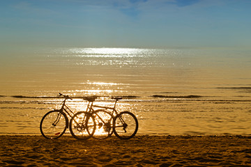 Two bicycles  silhouettes standing on the sand near the water edge on the beach.  Beautiful  shiny and glow water of the sea on the background. Sport and recreation concept.