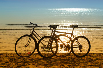 Two bicycles  silhouettes standing on the sand near the water edge on the beach.  Beautiful  shiny and glow water of the sea on the background. Sport and recreation concept.