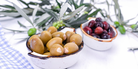 Traditional appetizers, green and red olives from Greek cuisine. White wood background. Fresh branches of olives. Copy space.