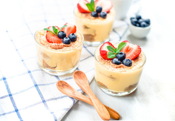 homemade, exquisite dessert tiramisu in glasses decorated with strawberry, blueberry, mint on white wooden table, top view.