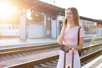 attractive girl is standing with luggage at the station and waiting for the train, the student goes on a trip, she goes on the platform and talks on the phone