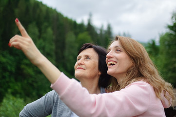 Happy senior mother and adult daughter are travelling and posing together over landscape of forest and mountains, young girl is pointing at something. Concept of travelling with family