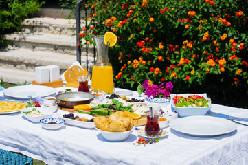 A breakfast table composed of cheese, orange juice, fried egg and various marmalades, placed in front of flowers at a garden