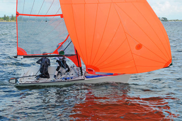 Two children sailing a racing dinghy with a large fully deployed orange coloured spinnaker.