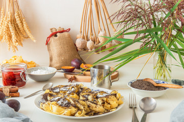 Sweet pasta dessert, noodles with poppy seeds, plum compote from fresh plums, dark background, white wooden table decorated with fresh flowers. Dry whole poppy plant in background.
