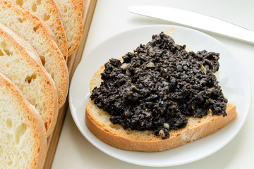 Tapenade sandwich on a sauser and sliced wheat bread on a wooden chopping board over white kitchen table. Homemade black olive paste with anchovies, garlic and olive oil.