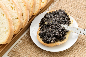 Tapenade sandwich and table knife on a sauser and sliced wheat bread on a burlap table napkin. Homemade black olive paste with anchovies, garlic and olive oil.