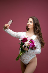 Beautiful pregnant woman in lace white lingerie body suit makes a selfie with a bouquet of flowers peony