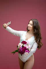 Beautiful pregnant woman in lace white lingerie body suit makes a selfie with a bouquet of flowers peony