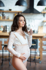 Beautiful pregnant woman with long hair in a lace jumper drinking a glass of water in the kitchen