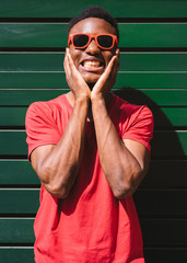 Portrait of young black man dressed with red t-shirt and sunglasses laughing on green background.