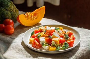 Vegetarian salad with broccoli, tomatoes, feta cheese, and pumpkin on white ceramic plate on linen textile, side view, hard light.