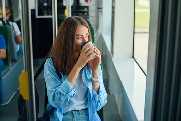 Beautiful young woman drinks delicious coffee in city bus or tram. Concept of public transport.