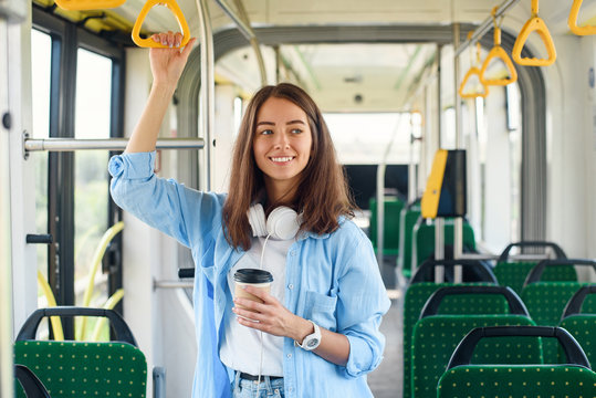 Stylish woman in blue shirt enjoying trip in the modern tram or bus, stands with cup of coffee in the public transport.