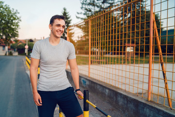 Portrait of young man standing on the street by the fence of the high school yard wearing gray t shirt and shorts in summer day