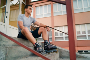 Portrait of young man in roller blade skates sitting on the stairs of the high school wearing gray t shirt and shorts in summer day