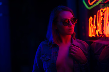 Fototapeta na wymiar Sexy portrait of a young girl in sunglasses and with in the night city, with creative light on the background of neon lamps. Night clubs, parties, strip business, night life. Banner, Horizontal photo