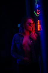 Fototapeta na wymiar Sexy portrait of a young girl with an open bust in sunglasses and with in the night city, with creative light on the background of neon lamps.Night clubs, parties, strip business, night life