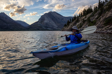 Fototapeta na wymiar Adventurous Man Kayaking in Glacier Lake surrounded by the beautiful Canadian Rocky Mountains during a cloudy summer sunset. Taken in Upper Waterton Lake, Alberta, Canada.