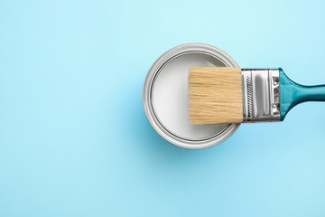 Open can with white paint and brush on blue background, top view. Space for text