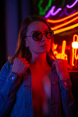 Sexy portrait of a young girl with an open bust in sunglasses and with in the night city, with creative light on the background of neon lamps.Night clubs, parties, strip business, night life.Vertical