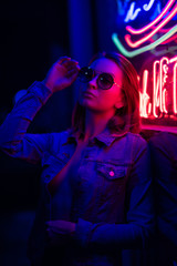 Fototapeta na wymiar Sexy portrait of a young girl with an open bust in sunglasses and with in the night city, with creative light on the background of neon lamps.Night clubs, parties, strip business, night life