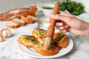 Woman brushing slices of delicious toasted bread with garlic and herbs on white marble table, closeup