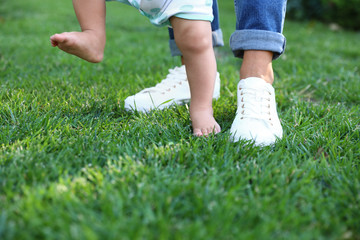 Cute little baby learning to walk with his nanny on green grass outdoors, closeup