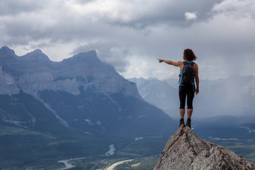 Obraz premium Adventurous Caucasian Girl pointing a finger on top of a rocky mountain during a cloudy and rainy day. Taken from Mt Lady MacDonald, Canmore, Alberta, Canada.
