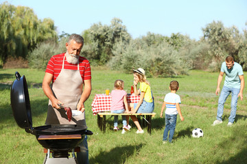 Happy senior man with meat at barbecue grill and his family having picnic in park