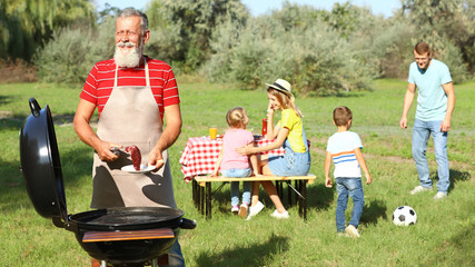 Happy senior man with meat at barbecue grill and his family having picnic in park