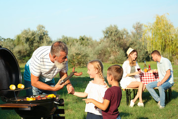 Grandfather with little kids cooking food on barbecue grill and their family in park