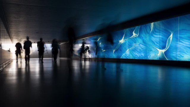 Timelapse of unrecognisable peoples silhouettes walking by in front of a blue illuminated glass wall. Pedestrian underpass
