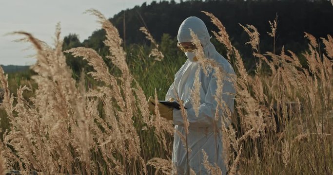 Ecological specialist in white protective costume staying at radioactive swamp in polluted landscape measuring contamination. End of civilization. Post-apocalypse.