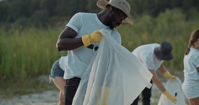 Young african man with friendly team of volunteers cleaning down polluted landscape in beautiful nature at lake. Ecology and environment.