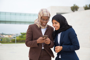 Excited happy office employees looking at smartphone screen outside. Muslim business women in hijabs standing outdoors, using mobile phones together and laughing. Digital technology concept