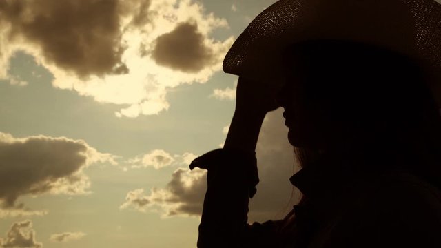 long haired girl silhouette puts on cowboy hat against grey clouds hiding bright sun slow motion close view