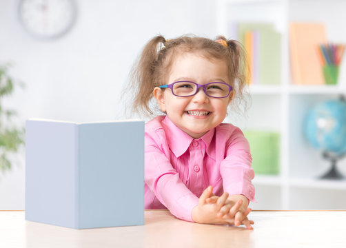 Happy kid girl in eyeglasses reading with standing book on table