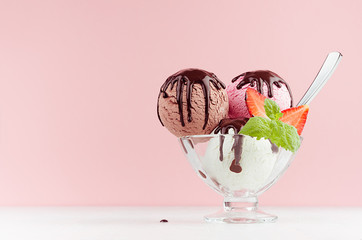 Set of different refreshing classic creamy ice cream balls in bowl with chocolate sauce, mint, spoon, strawberry on white wooden table, pink background.