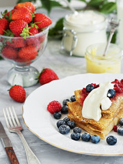 Pancakes with jam on  white plate, around the pancakes on a plate are fresh berries. Next to a glass of milk and sour cream. Festive food. The table is decorated with an ivy branch.