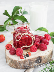 A jar of jam is located on a wooden plate, with fresh berries lying around. In the background is a glass of milk. The table is decorated with ivy and thuja branches. Festive food. Gray background. 