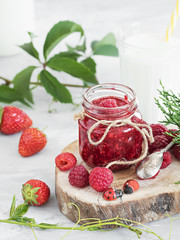A jar of jam is located on a wooden plate, with fresh berries lying around. In the background is a glass of milk. The table is decorated with ivy and thuja branches. Festive food. Gray background. 