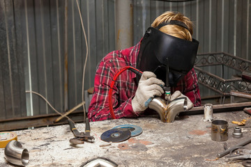 Young guy welder in a checkered red shirt welds a stainless steel pipe using agronomic welding to protect his eyes with a mask in an iron workshop. Modern welding methods.